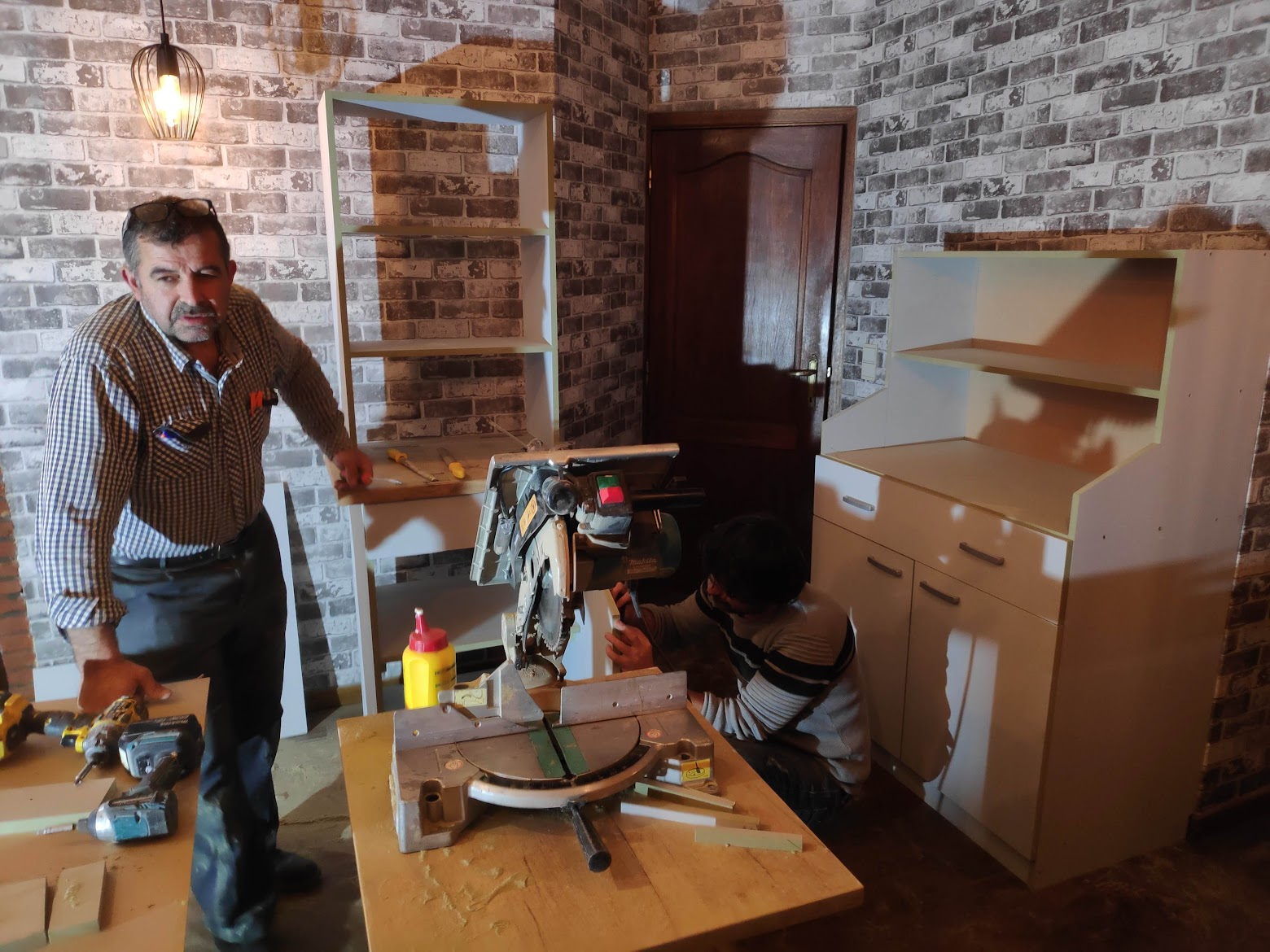 Bouwproces van de buffetkast op de bovenverdieping met zaagmachine, onderdelen en twee makers.